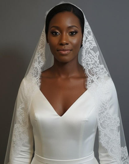 May include: A bride in a white satin wedding dress with a V-neck and long sleeves. The dress is complemented by a delicate lace veil. The veil and sleeves have detailed lace. The background is a plain grey.