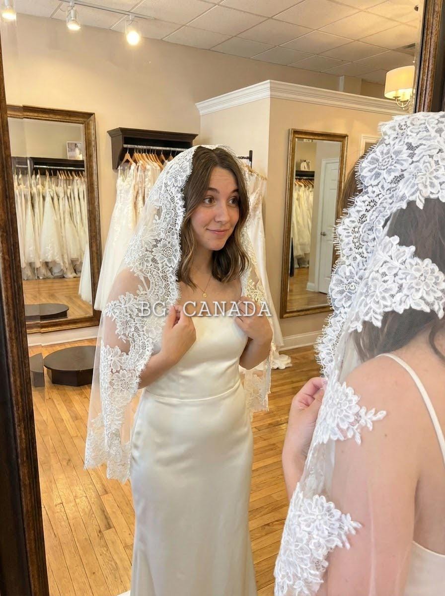 May include: A bride in a satin wedding dress and a white lace veil. The veil has detailed floral lace edging. The bride is in a bridal shop, standing in front of a mirror.